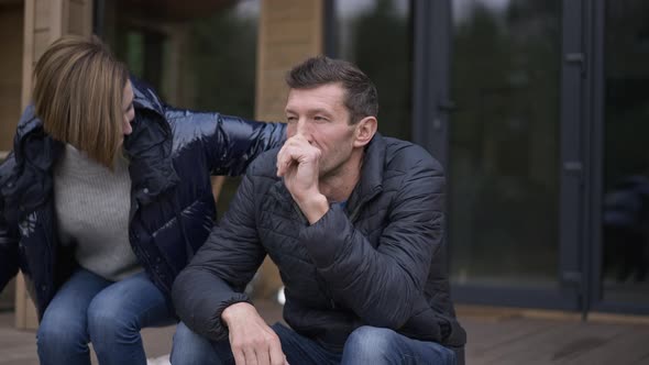 Thoughtful Man Sitting on Wooden Porch at Home As Woman Coming Hugging Spouse alt