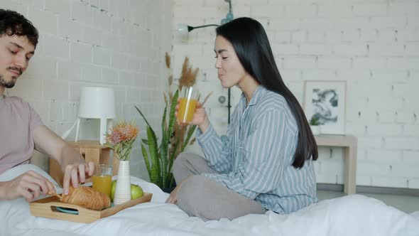 Girl and Guy Eating Breakfast and Talking Smiling in Bed in Apartment alt