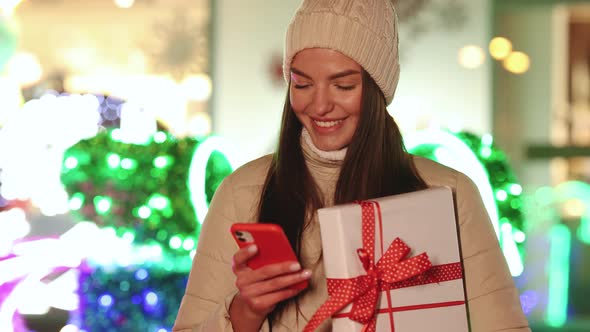 Portrait of Happy Caucasian Woman Standing Outdoor With Christmas Gift and Texting on Smartphone alt