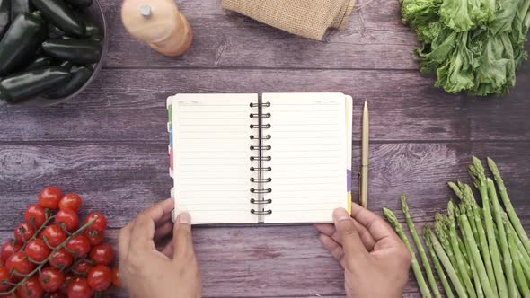 Top View of Man's Hand Turning Pages of Notepad with Fresh Vegetable on Table alt