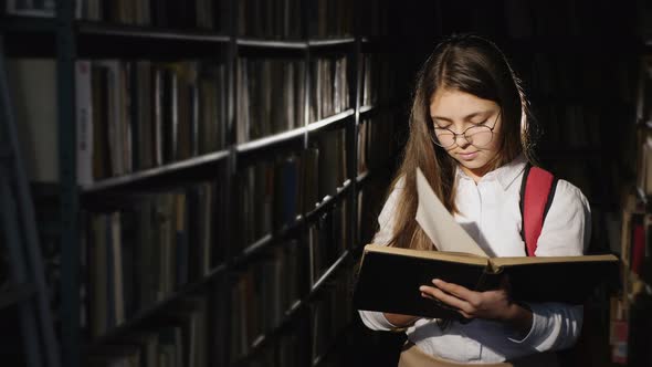 Thoughtful Schoolgirl Reads a Book Standing in the Library