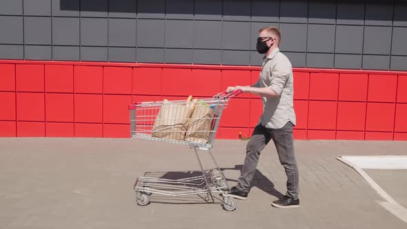 Man in Face Mask Pushing Cart of Groceries Outdoors alt