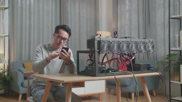 Asian Man Using Smartphone And Looking At The Mining Rig For Mining Cryptocurrency alt