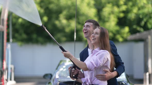 Young Couple Laughing Turning on High Pressure Washer Standing at Car Wash Service Outdoors on Sunny alt