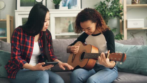 Asian Woman Is Teaching Her African-American Friend To Play the Guitar During Leisure Time at Home alt