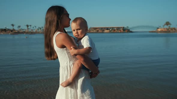 Long Hair Teenage Girl Walking Along Sunny Seaside with Baby Boy on Hands. alt