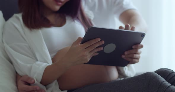 Asian husband and pregnant wife spending time together in the bedroom and using a tablet.