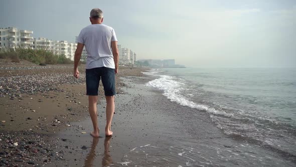 A Slender Elderly Grayhaired Man Slowly Walks Barefoot on a Deserted Beach in the Running Waves in