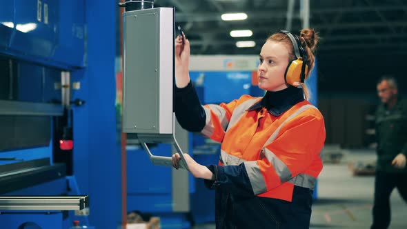 Female Worker in Ear Protectors Uses Portable Screen to Operate a Machine alt