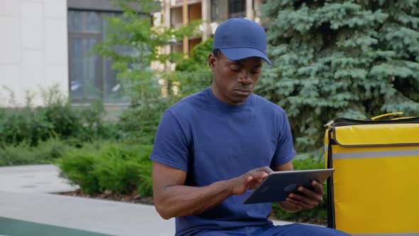 Afroamerican Man Courier Delivery Sitting on Bench with Backpack Uses Tablet alt
