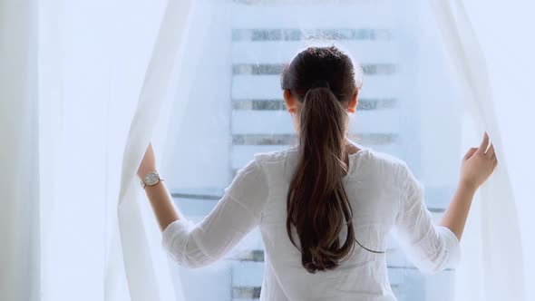 Young asian woman relax open curtains at the window looking outside. alt