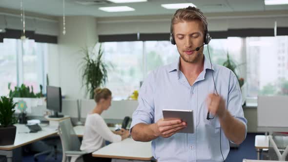 Young Handsome Businessman in Headset Holding Tablet Showing Thumb Up Coworker Working on Background alt