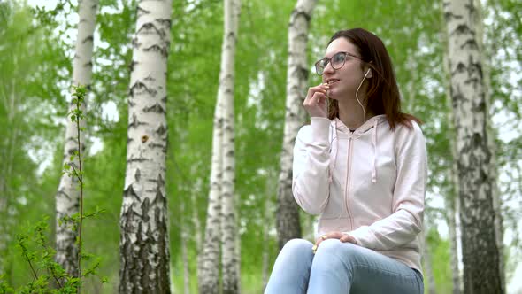 A Young Woman in Nature Is Talking on the Phone Through Headphones. A Girl Sits on a Stump in a alt