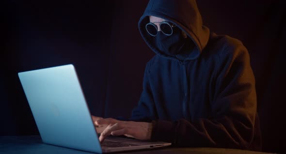 A hacker with a closed face sits at a laptop in a dark room alt