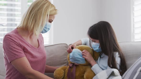 Girl putting face mask on her teddy bear toy at home alt