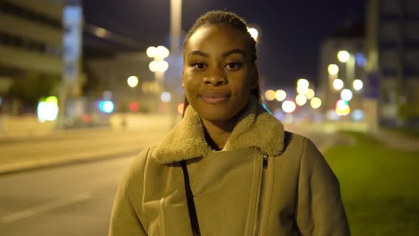 A Young Black Woman Smiles at the Camera in a Street in an Urban Area at Night alt