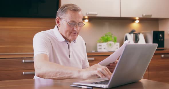 Happy Elderly Man Sitting on Chair in Kitchen and Buying with Laptop Use Online Website Voucher and alt