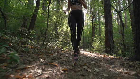 Female Jogger in Sports Outfit and Sports Shoes Running Down a Forest Path, Carrying Water Bottle alt