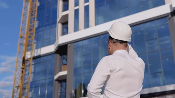 An Architect Engineer in a White Shirt and Helmet Stands with His Back to the Camera Against the alt