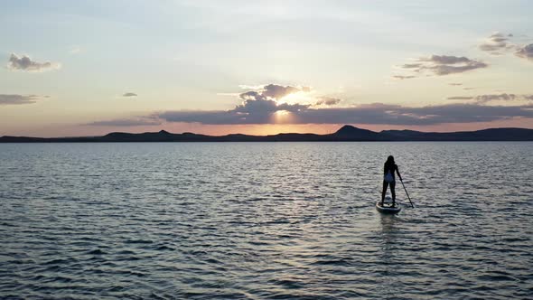 A Young Woman is Rowing an Oar Standing on a Board in the Lake at Sunset