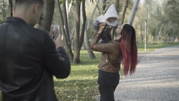 Young Caucasian Man Taking Photos of Smiling Beautiful Woman Playing with Infant in Sunny Park alt