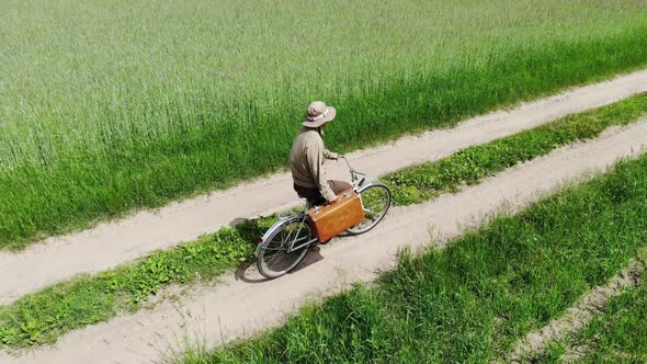 Man Riding on Bicycle By Country Road in a Green Field, Holding Suitcase in a Hand, Funny Travel