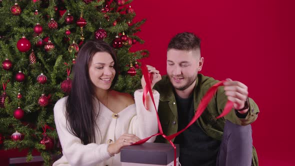 Young Loving Couple Opening Long Awaited New Year Gift Sitting Near Xmas Tree