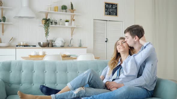 Man Hugs Lady Sitting on Sofa and Kissing in Kitchen alt