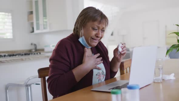 Sick african american senior woman with face mask using laptop at home, with medication on table alt