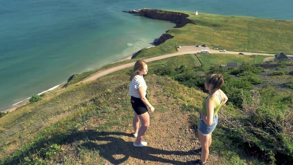 Two women talking as they stand overlooking the sea and a park far below. Sunny, breezy coastal loca alt