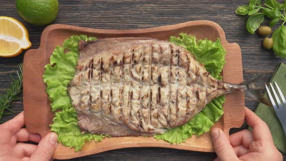 Hands are Putting Plate with Grilled White Mackerel Fish on a Dark Wooden Table alt