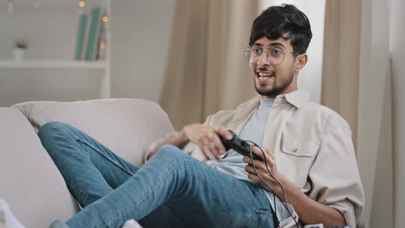 Arab Guy Bearded Man with Glasses Lying on Couch at Home Playing Console Online Video Game alt