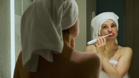 Woman with White Bath Towel on Head Brushing Her Teeth Looking in the Mirror alt