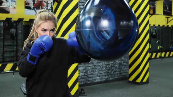 Young Female Athlete in Blue Boxing Gloves Hits a Pear