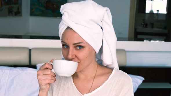 Cheerful Girl in a Bathrobe and Towel Drinks Coffee While Sitting on the Bed Looking at the Camera