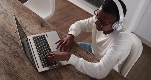 Young latin man typing on computer laptop while wearing headphones at home alt