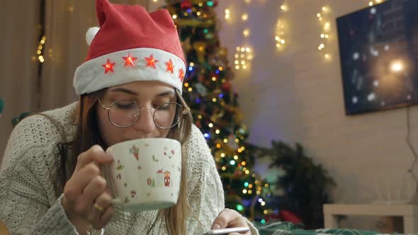 A Woman in a Red Santa Claus Hat Lies on a Sofa Looks at the Screen of the Phone and Drinks Hot