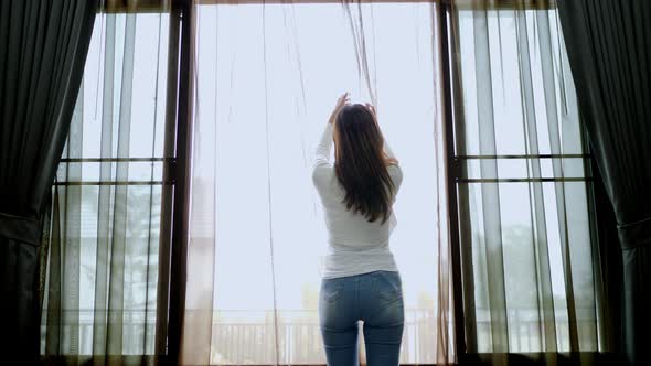 Young Woman opens curtain and looks out of window alt