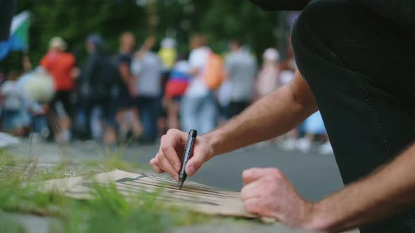 Male Protester Drawing Banner Sign in Hands with Marker in Rally Riot Crowd alt
