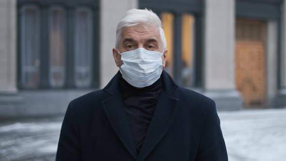 Old Graying Man in Coat and Covid 19 Face Mask Walks on Epidemic European Street alt