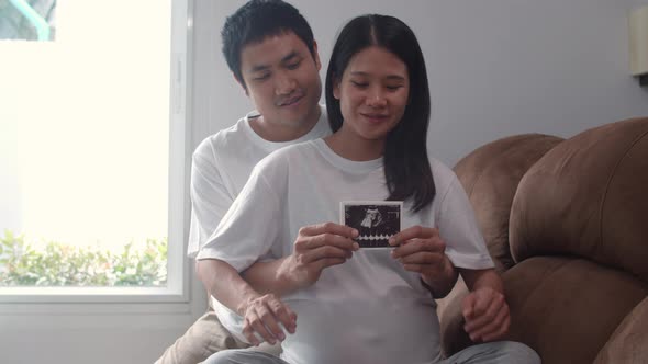 Young Asian Pregnant couple show and looking ultrasound photo baby in belly feeling happy smiling.