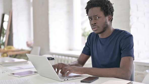 Shocked African Man Looking at Camera While Working on Laptop alt
