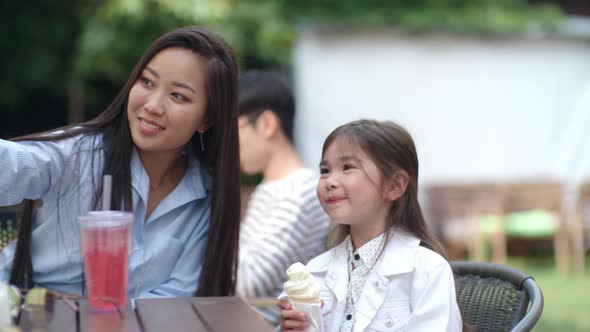 Asian Woman Taking Selfie with Little Girl in Outdoor Cafe alt