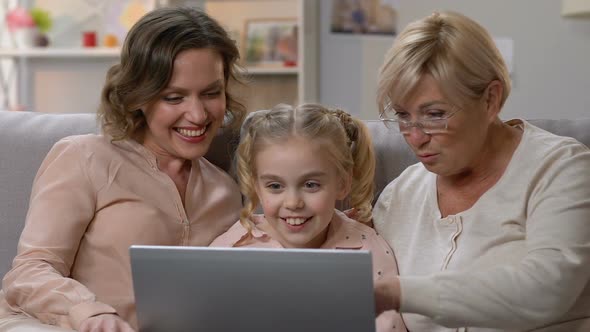Little Girl Showing Funny Video on Laptop to Mom and Granny, Humorous Show alt