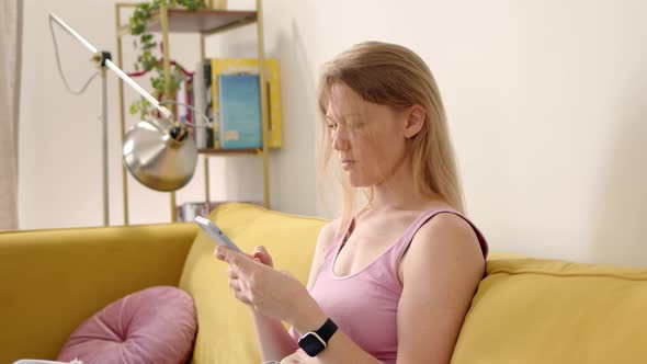 Blonde Sits on Yellow Sofa in Pink Top and Use Phone Typing Text alt
