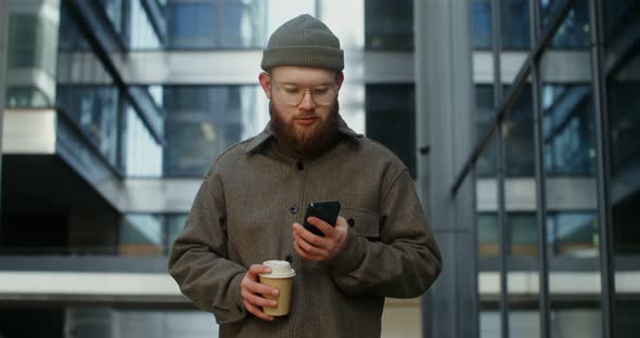 A Bearded Man is Typing on a Mobile Phone While Standing Near Office Building alt