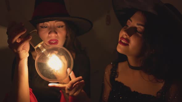 Two Colombian Ladies In Witch Hats Moving Hands Around A Lit Light Bulb In Dark Room alt