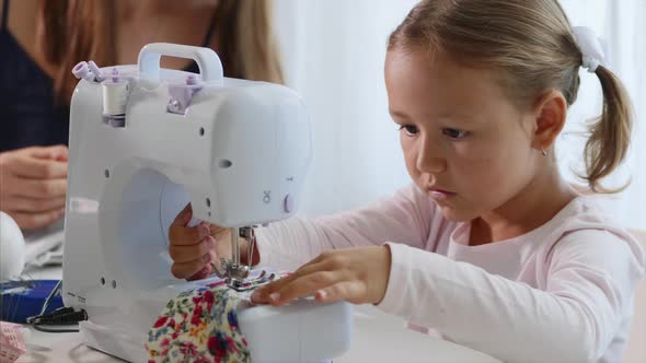 A Little Girl Is Sewing Fabric and Talking. Mom Is at the Background. Close-up