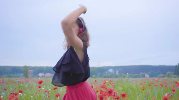Pretty Young Woman Wearing Headphones Listening To Music and Dancing in a Poppy Field Smiling alt