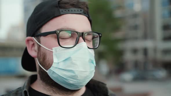 Portrait of Young Man in Glasses Cap and Medical Mask Looks Aside Thoughtfully alt